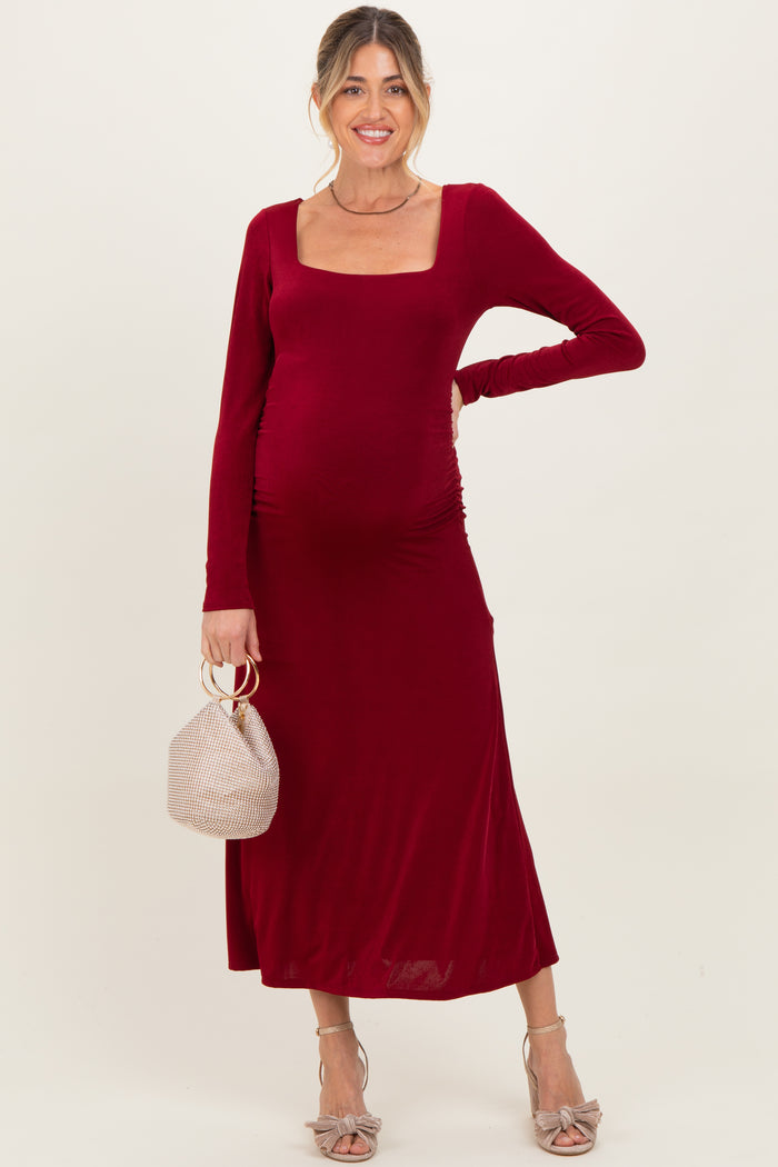 woman wearing a burgundy long sleeve maternity dress with a square neckline and gathered sides, holding a small bag