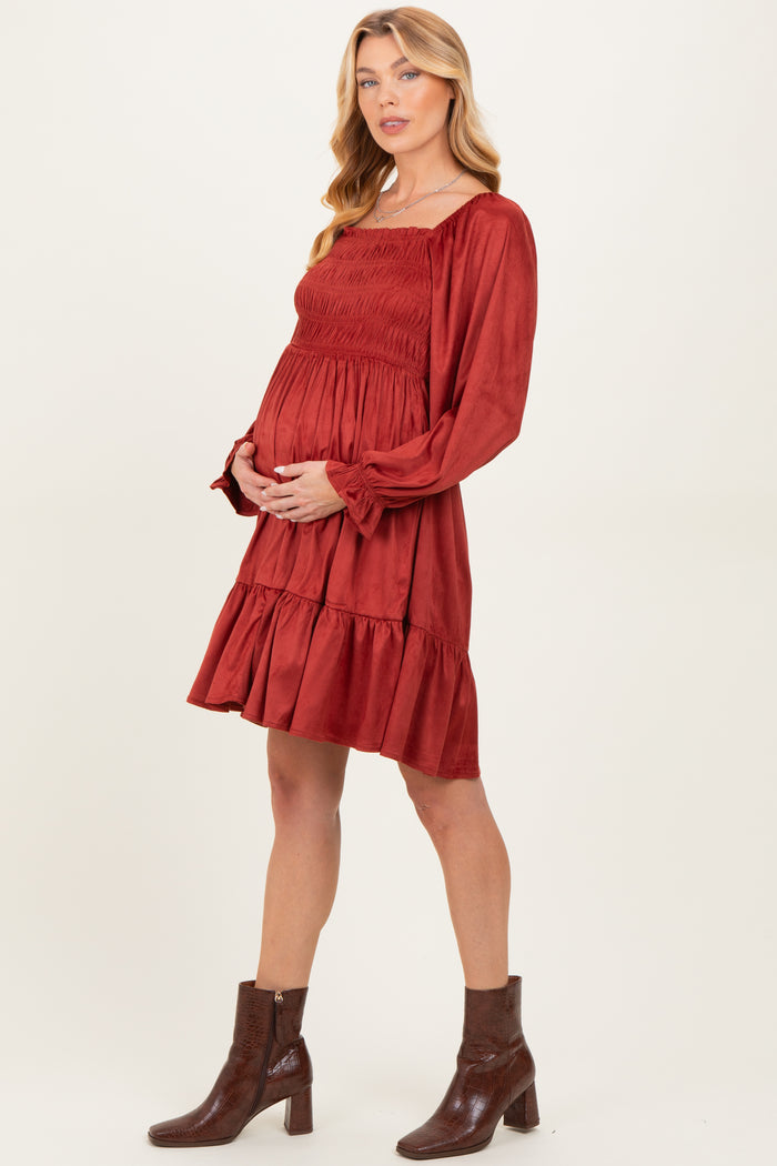 woman wearing a rust colored maternity dress with long sleeves, smocked bodice, and tiered skirt, holding her baby bump