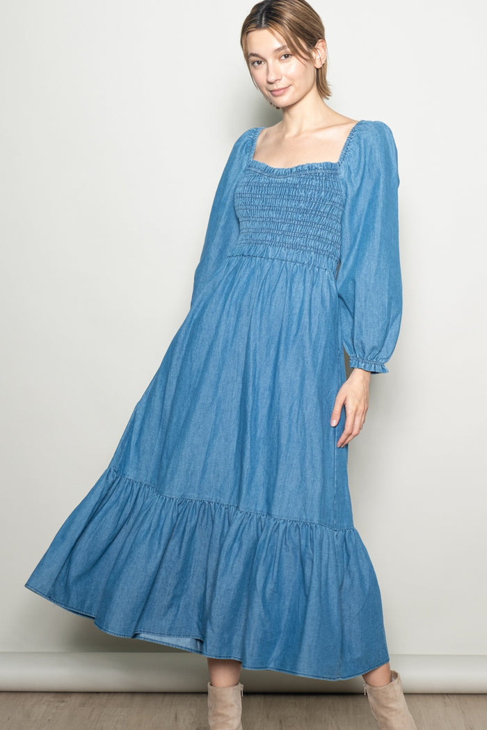 woman wearing a blue denim maxi dress with puff sleeves and a smocked bodice, standing against a neutral background