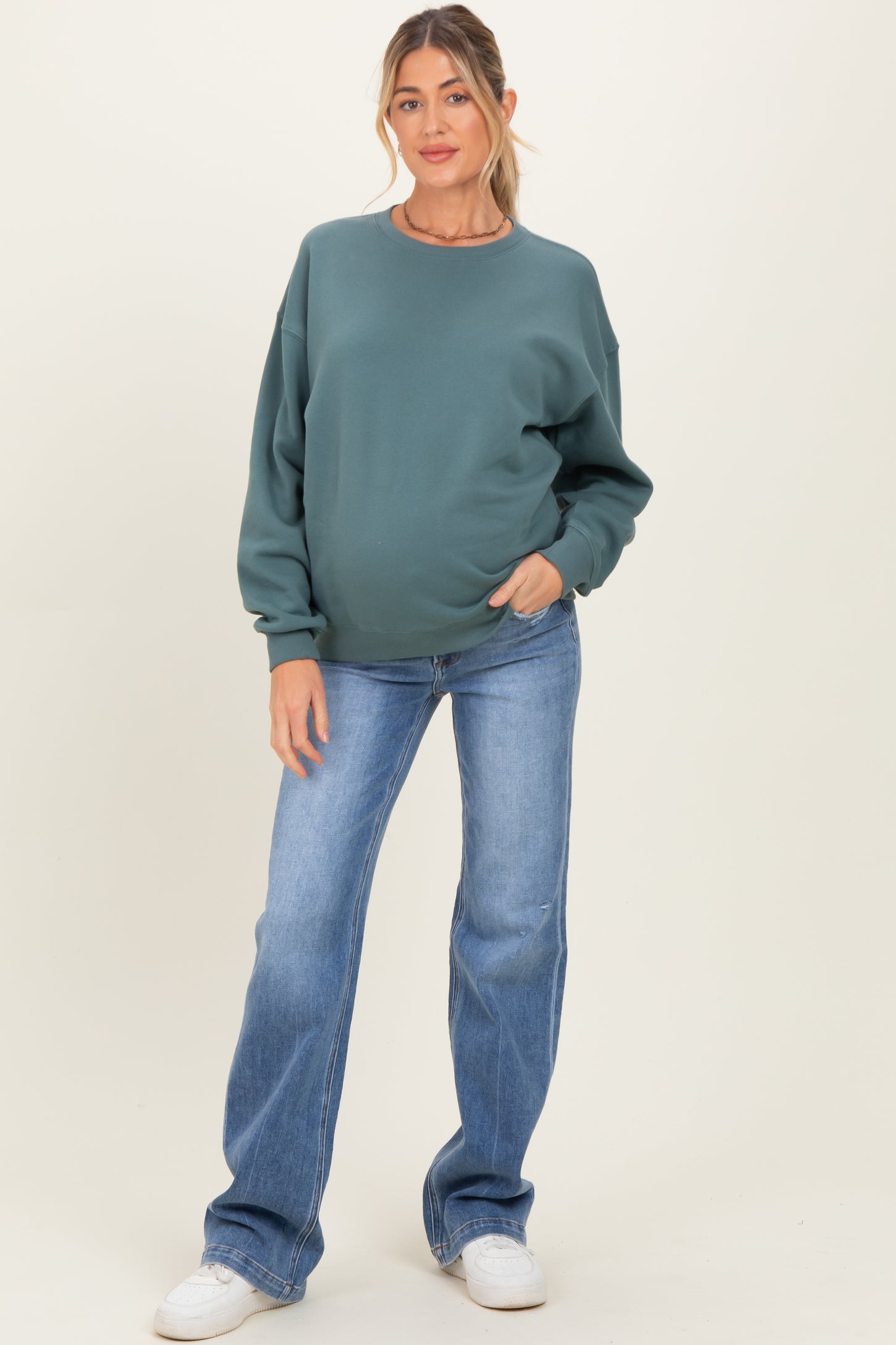 woman wearing a green oversized maternity sweatshirt and blue flared jeans, showing her baby bump