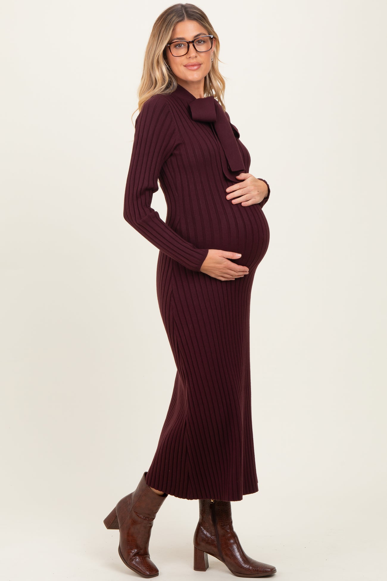woman wearing a burgundy ribbed maternity dress with a bow detail, gently cradling her baby bump