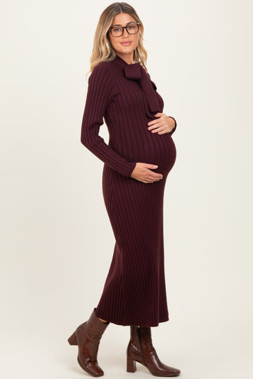 woman wearing a burgundy ribbed maternity dress with a bow detail, gently cradling her baby bump