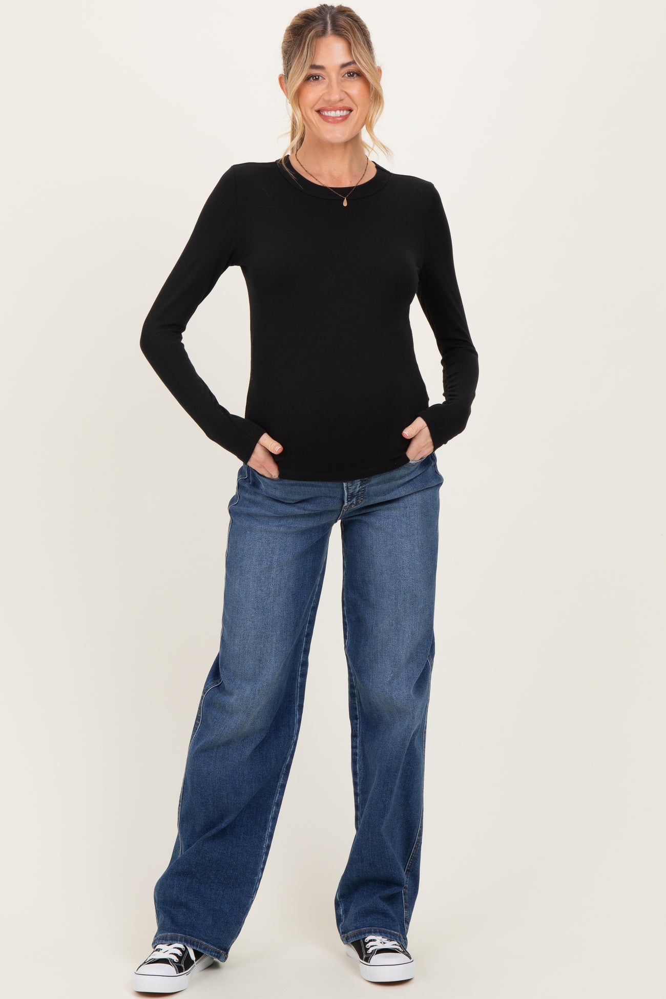 woman wearing a black long sleeve maternity top and loose blue jeans, smiling with hands in pockets