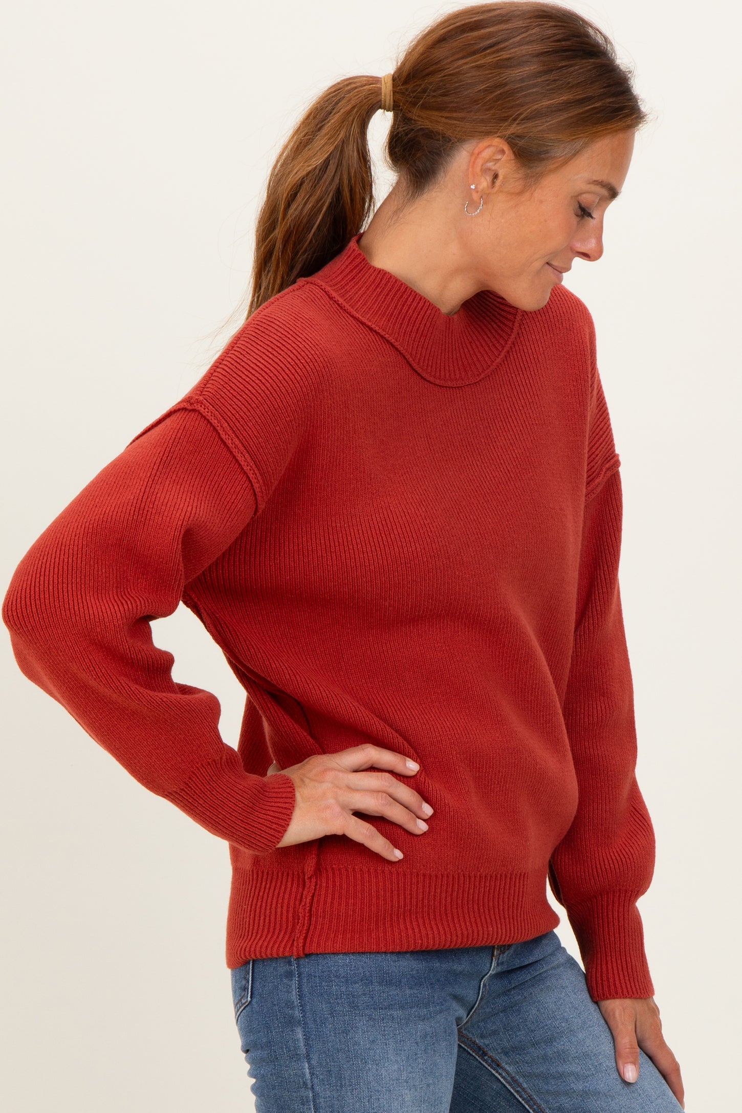 woman wearing a rust-colored oversized ribbed sweater with a high neckline and long sleeves, styled with blue jeans