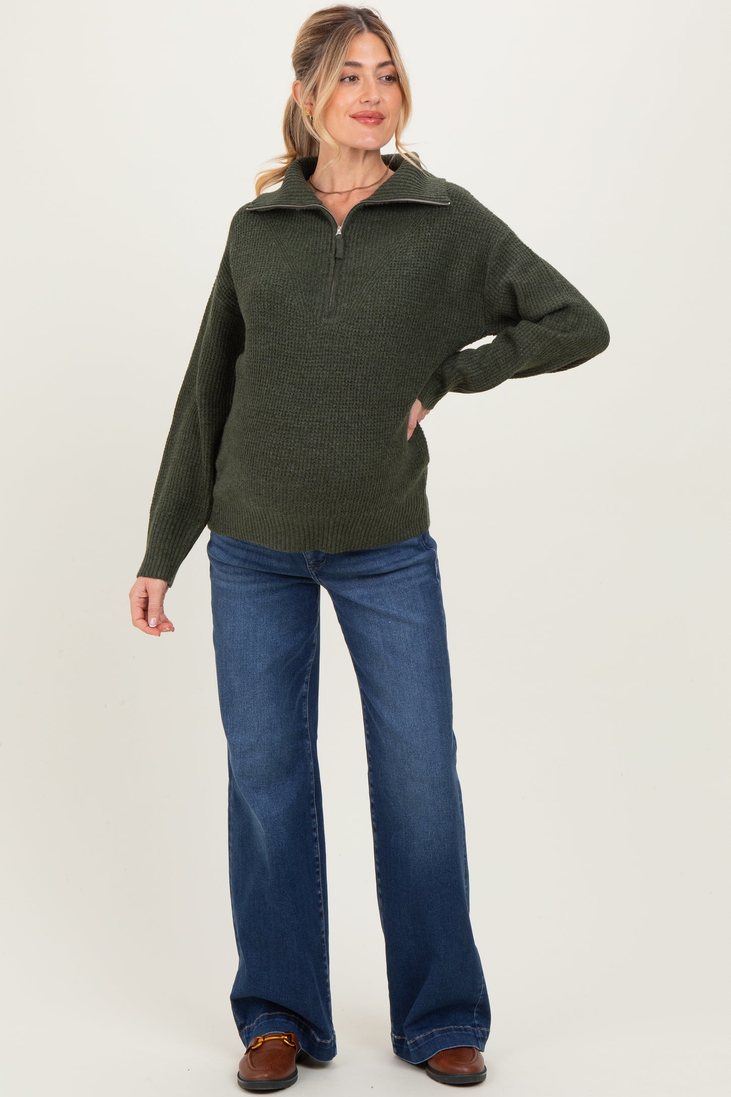 woman wearing a dark green maternity zip-up sweater and flared jeans, posing with a relaxed expression