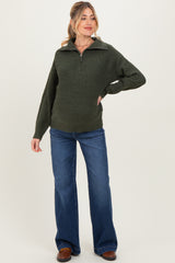 woman wearing a dark green maternity zip-up sweater and flared jeans, posing with a relaxed expression
