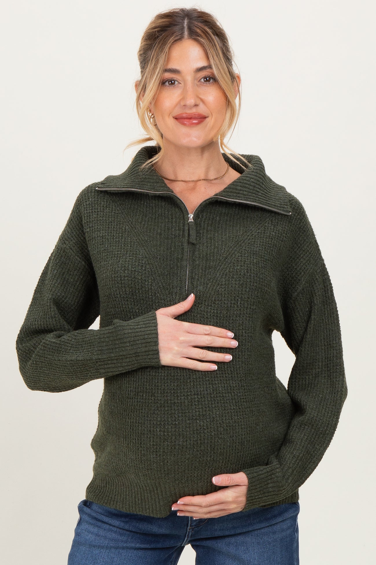woman wearing a dark green maternity sweater with a half-zip collar and holding her baby bump