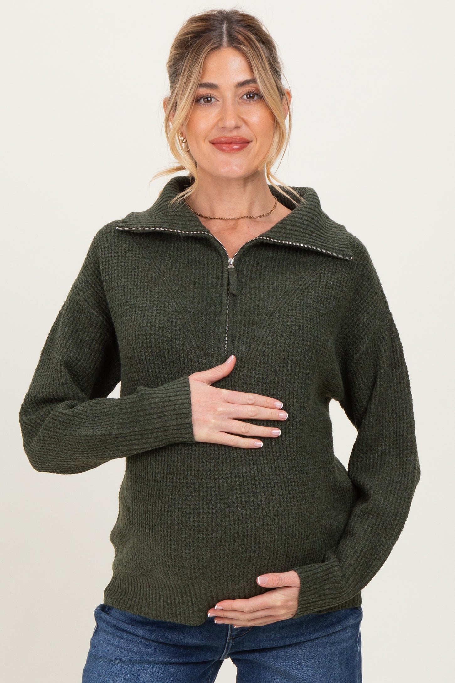 woman wearing a dark green maternity sweater with a half-zip collar and holding her baby bump