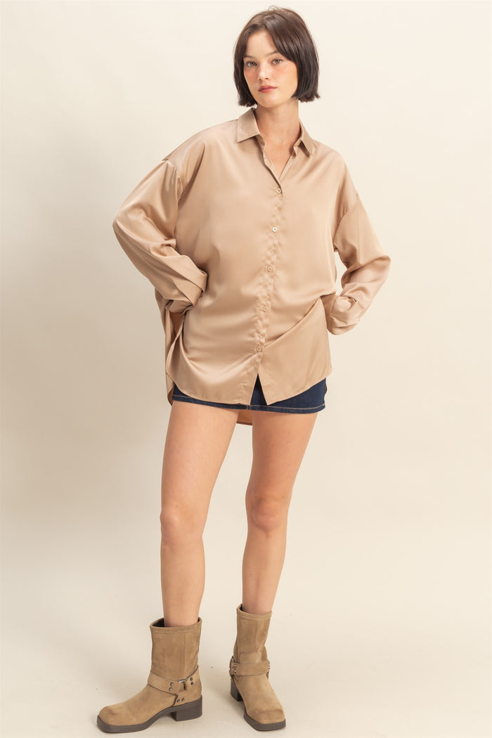 woman wearing a beige oversized satin button-up shirt with rolled sleeves and denim shorts, standing in a relaxed pose