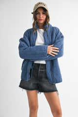 woman wearing a blue quilted jacket over a white t-shirt and black denim shorts, styled with a beige cap