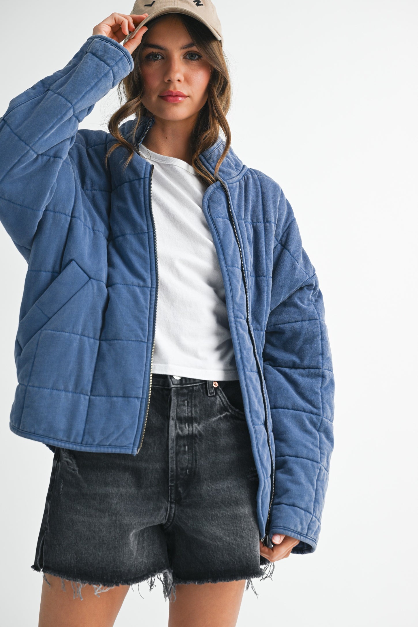 woman wearing a blue quilted jacket over a white t-shirt and black denim shorts, holding a beige cap
