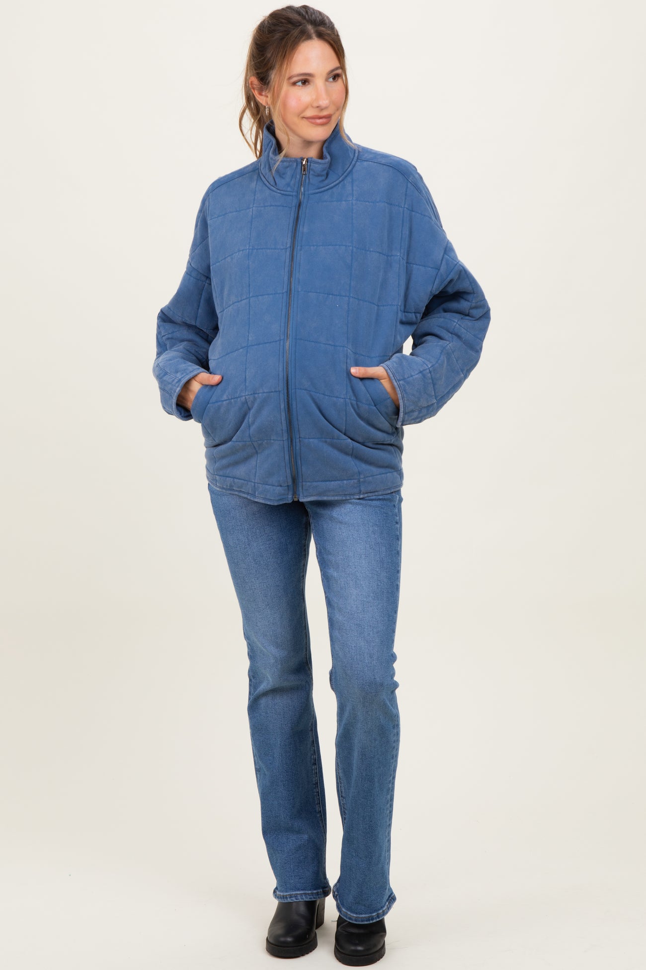 woman wearing a blue quilted maternity jacket with a zip front and pockets, paired with flared jeans and a visible baby bump