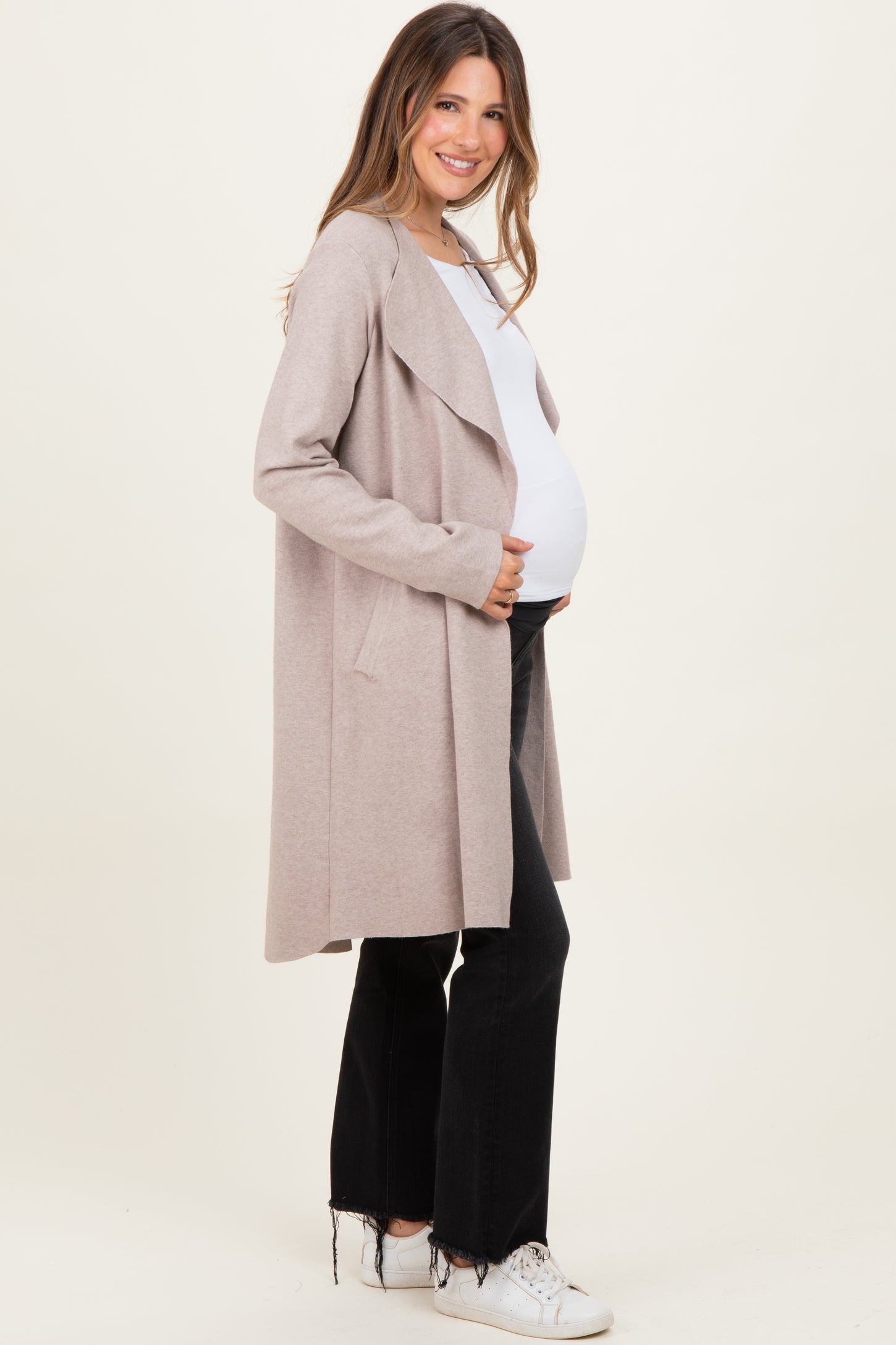 woman wearing a light beige maternity cardigan over a white top, holding her baby bump, paired with black jeans and white sneakers