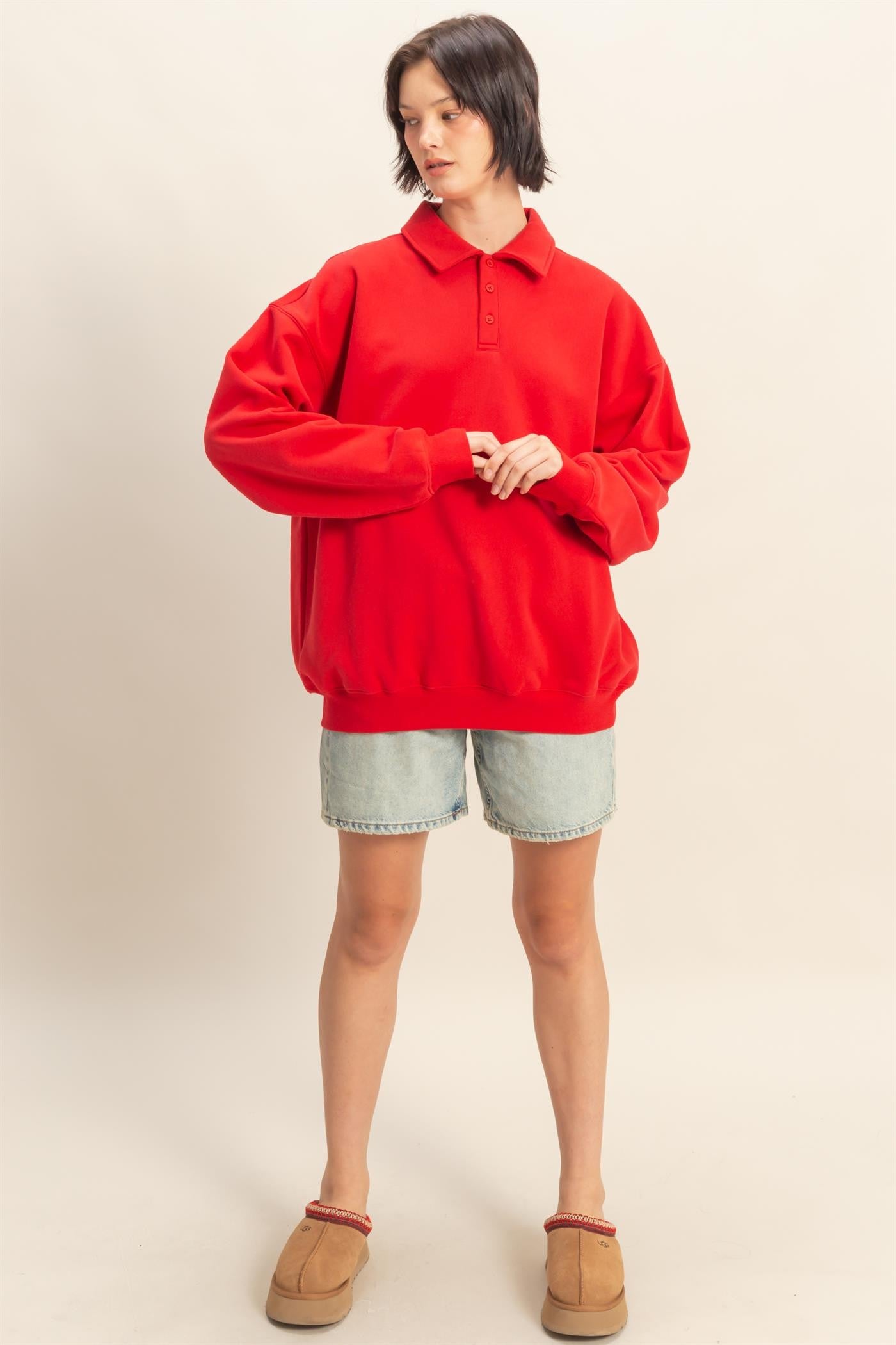 woman wearing a red oversized polo sweatshirt with long sleeves and denim shorts, standing in casual footwear