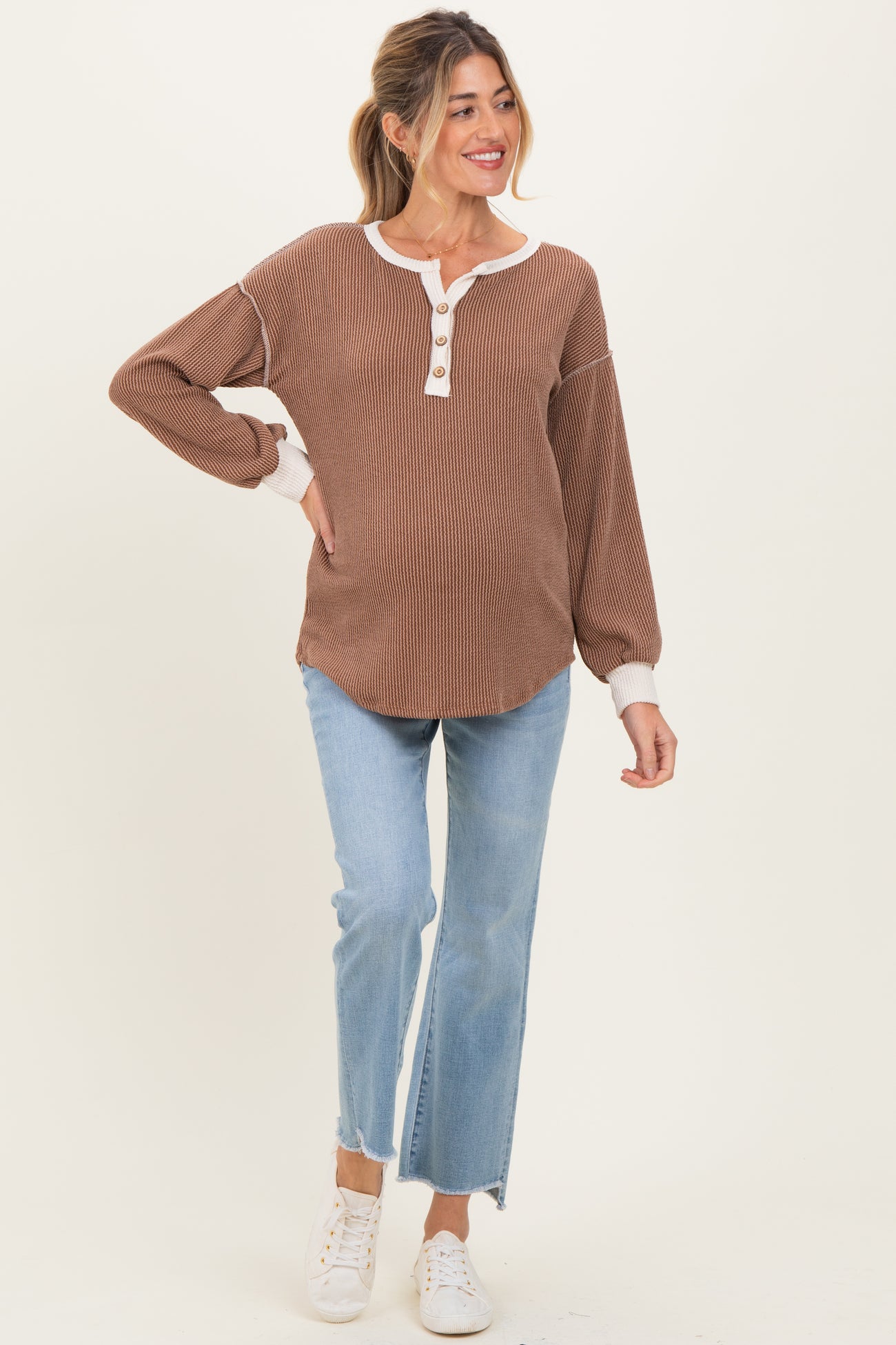 woman wearing a brown ribbed maternity top with white buttons and a white collar, paired with light blue jeans and white sneakers