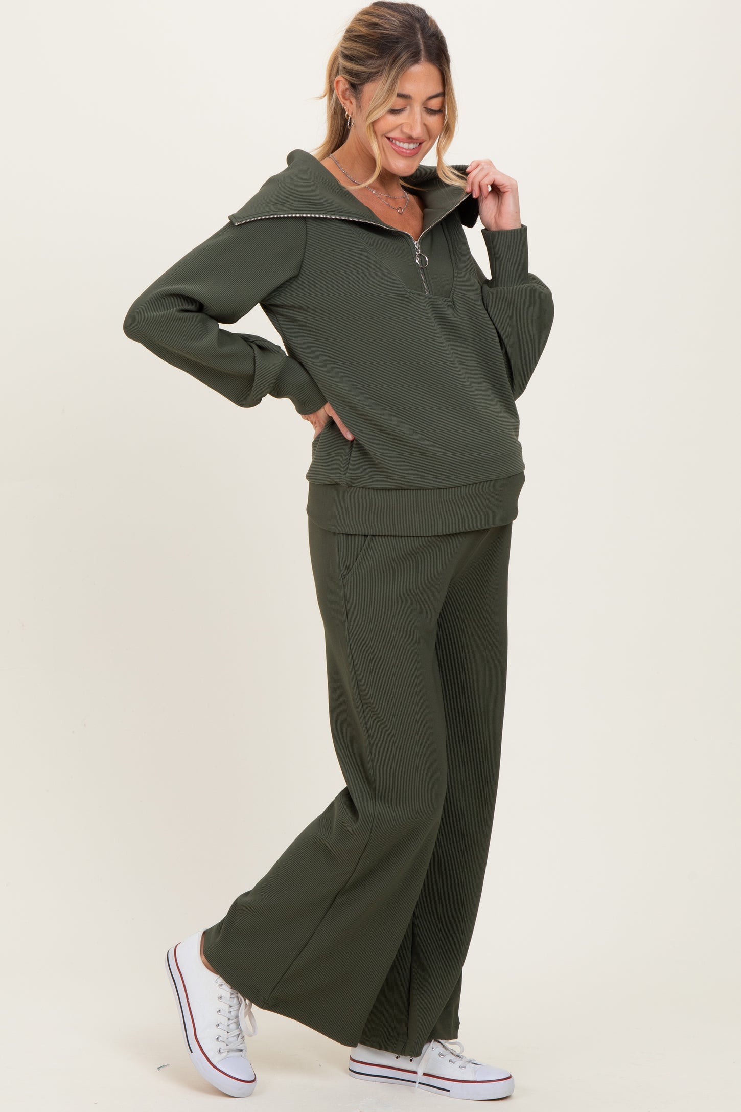 woman wearing a dark green maternity zip-up sweatshirt and matching wide-leg pants, styled with white sneakers
