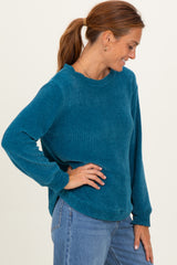 woman wearing a teal long sleeve textured sweater and blue jeans, smiling and posing sideways