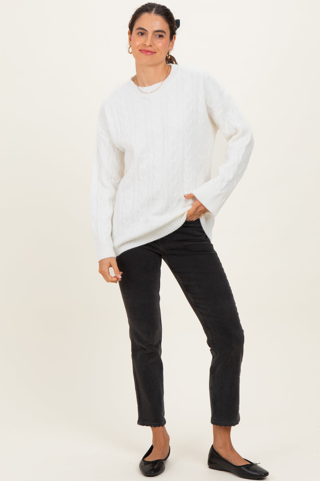 woman wearing a white cable knit sweater and black jeans, standing with one hand in pocket and smiling