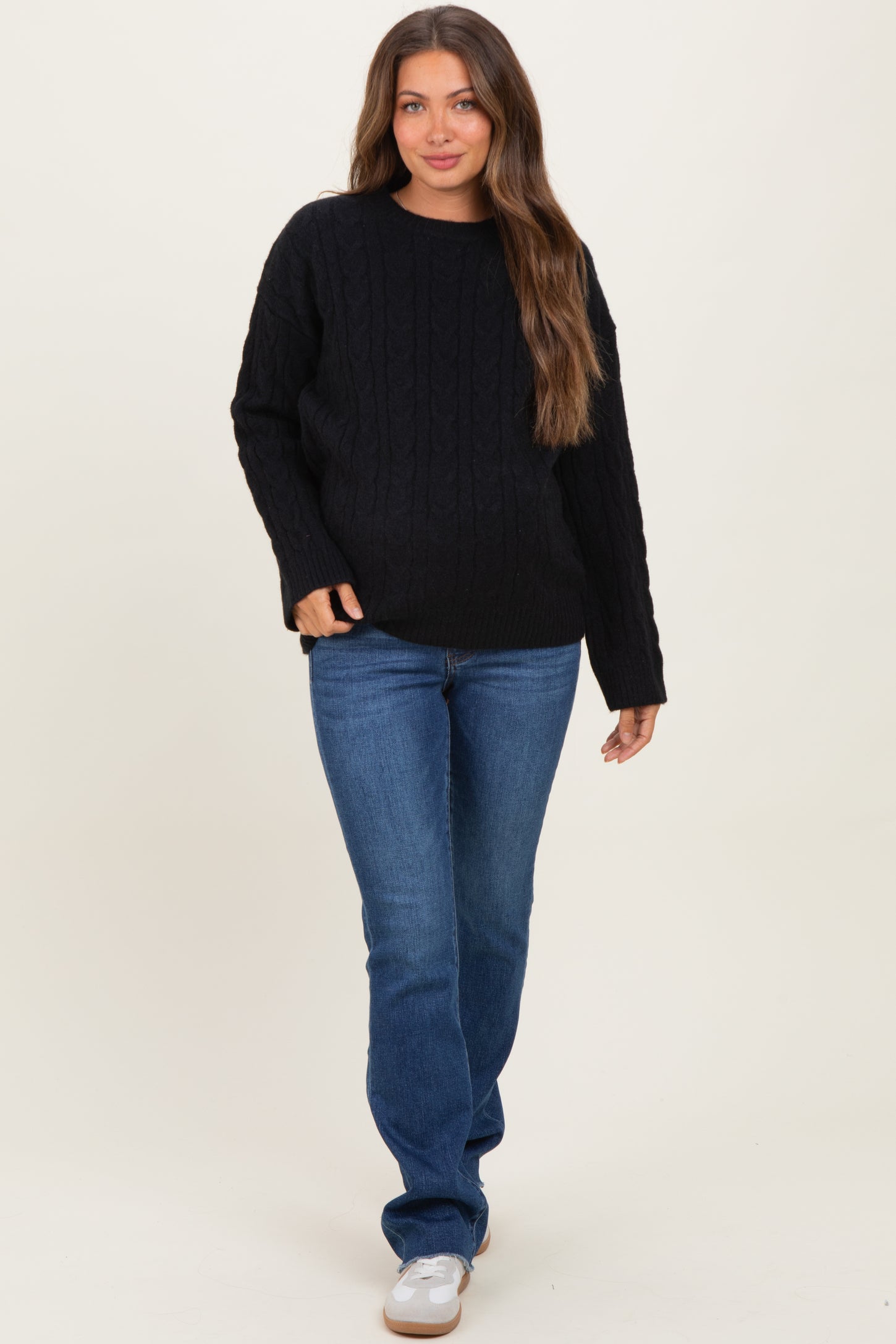 woman wearing a black cable knit maternity sweater and blue jeans, with long brown hair and a relaxed pose
