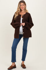 woman wearing a brown maternity cardigan over a white tank top and blue jeans, with a visible baby bump