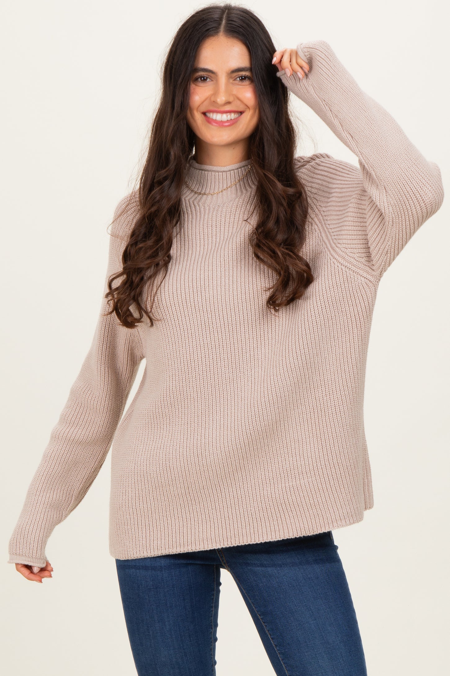 woman wearing a beige ribbed knit sweater with a high neck and long sleeves, smiling in front of a light background