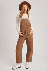 woman wearing brown overalls with a white long sleeve top and a brown hat, standing confidently against a neutral background
