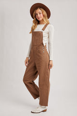 woman wearing a tan overall jumpsuit with side buttons and a long sleeve white top, accessorized with a brown hat