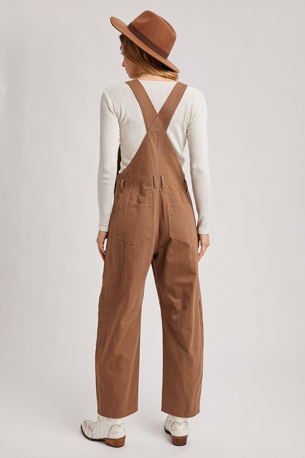 woman wearing a cream long sleeve top and brown denim overalls with wide legs and a brown hat