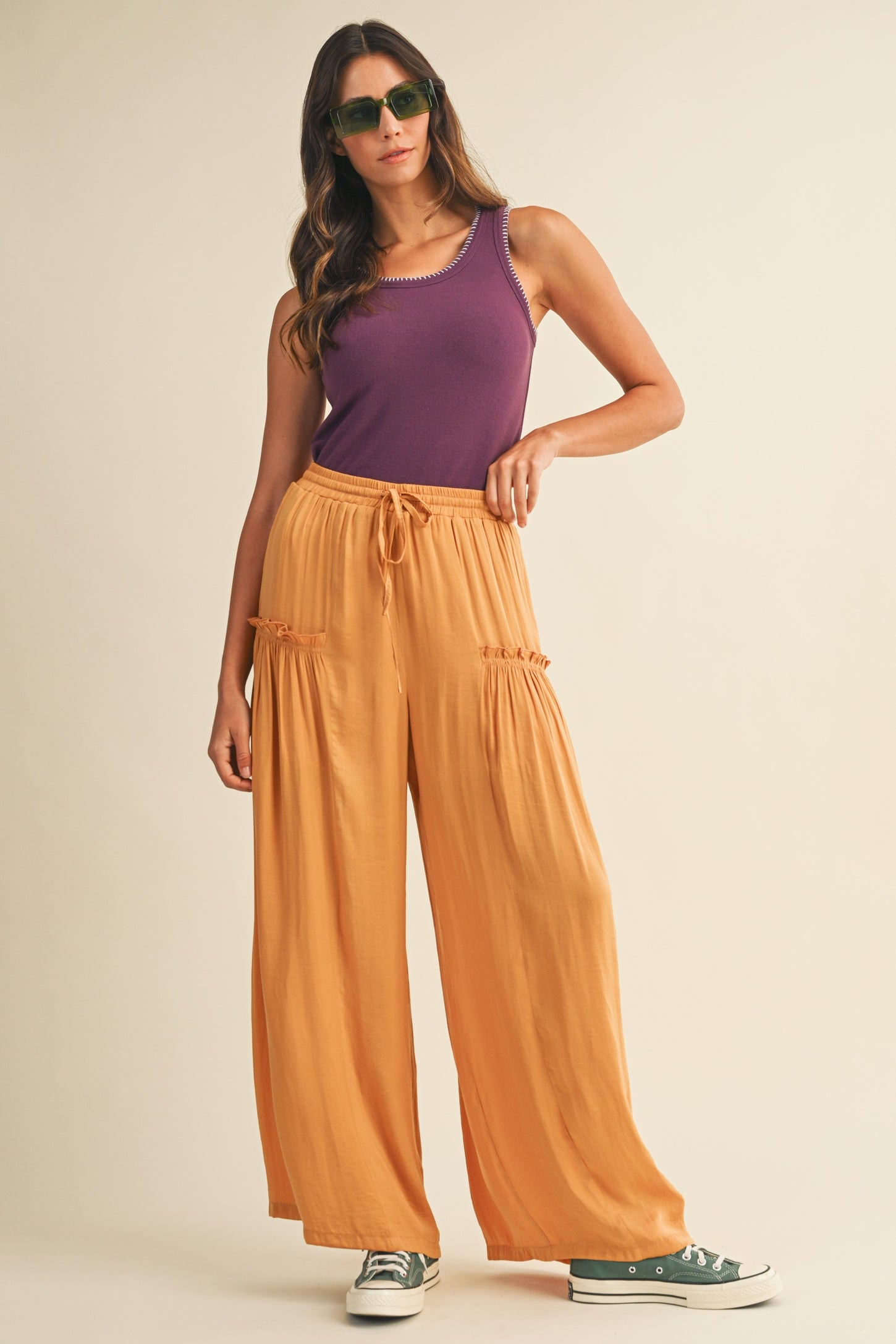 woman wearing a purple tank top and orange wide-leg pants, accessorized with green sunglasses and sneakers
