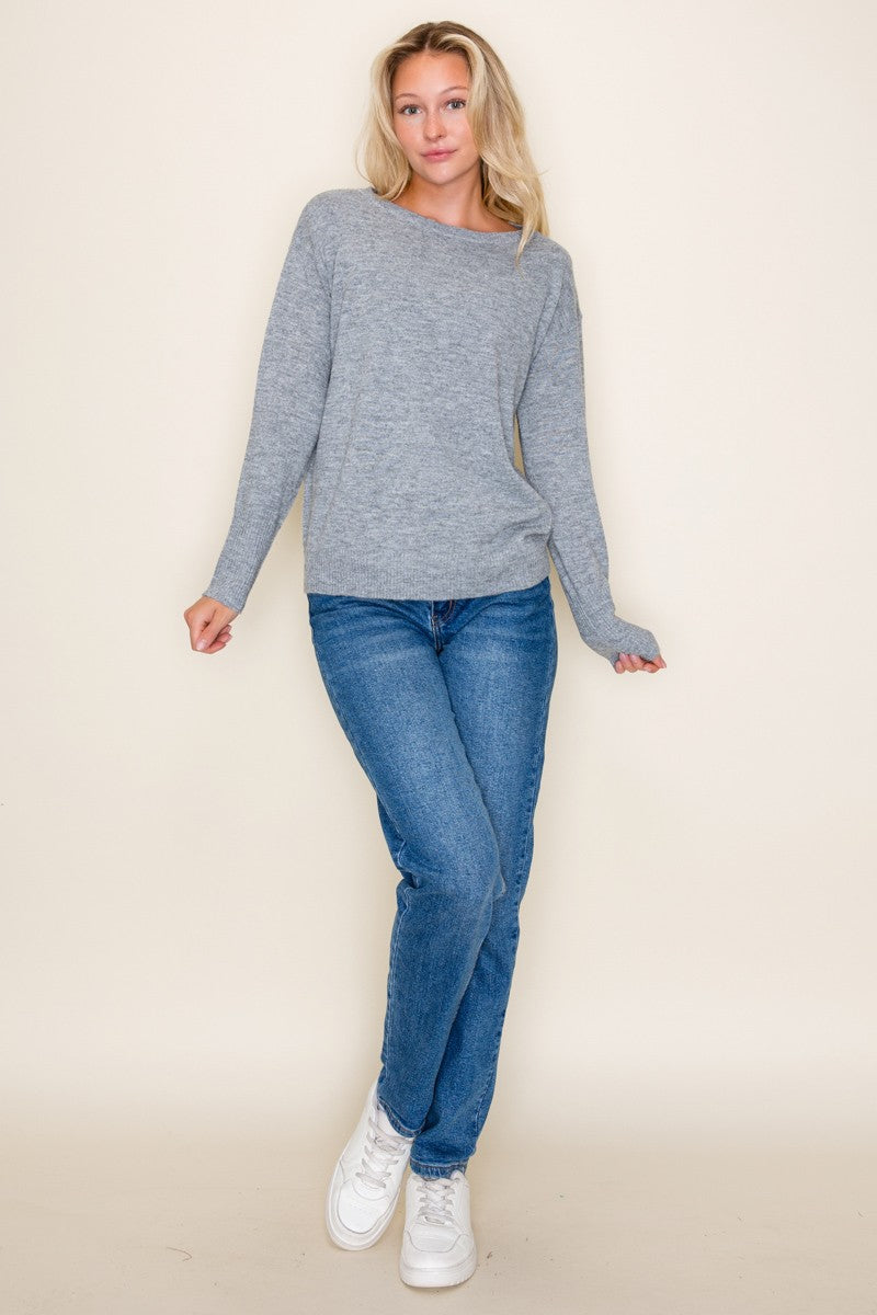 woman wearing a light gray long sleeve sweater paired with blue jeans and white sneakers standing against a neutral background