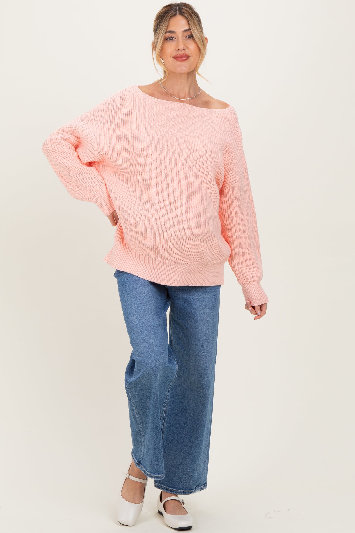 woman wearing a light pink oversized maternity sweater with off-shoulder detail and blue wide-leg jeans, showcasing her baby bump