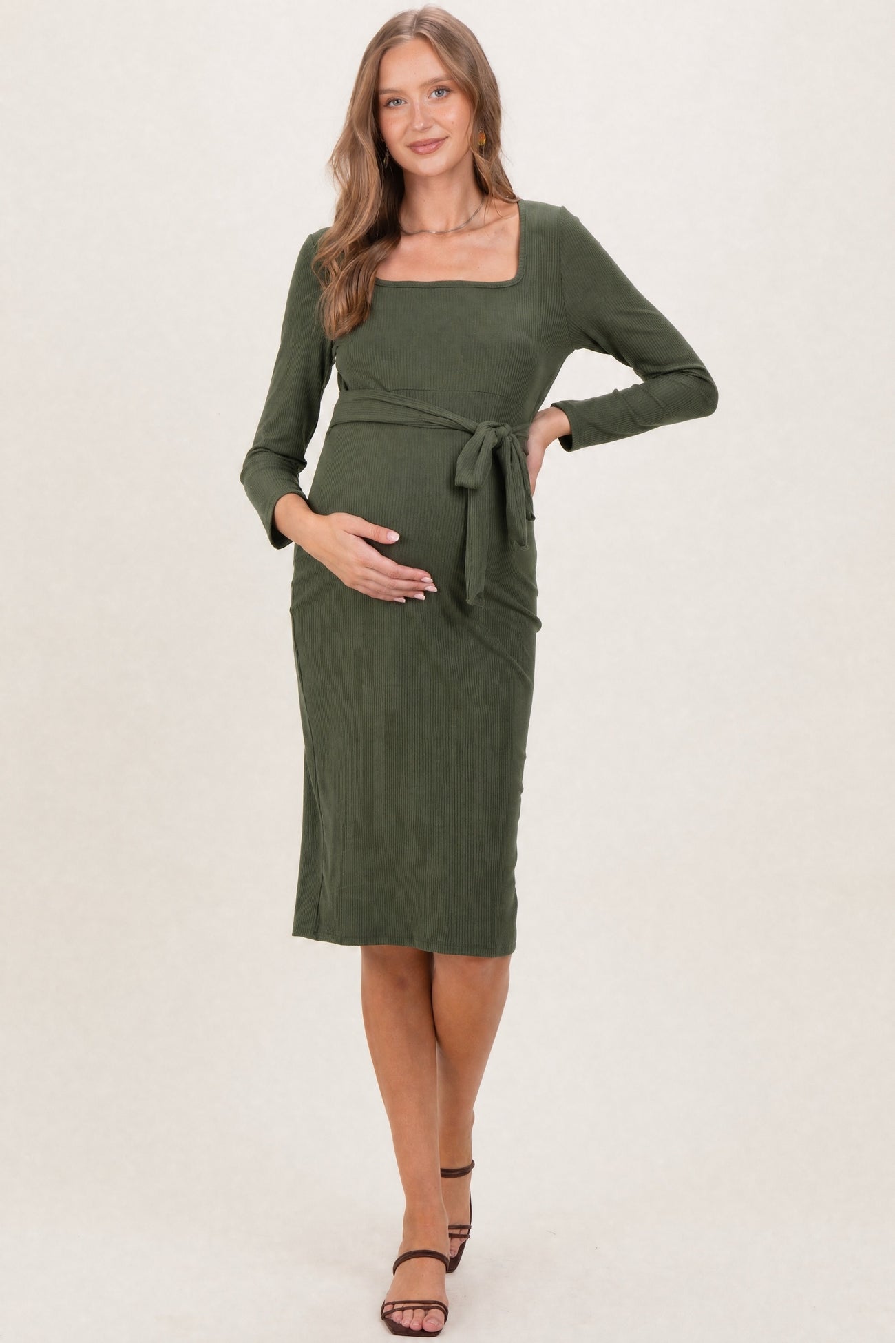 woman wearing a dark green ribbed maternity dress with a square neckline and tied waist, cradling her baby bump