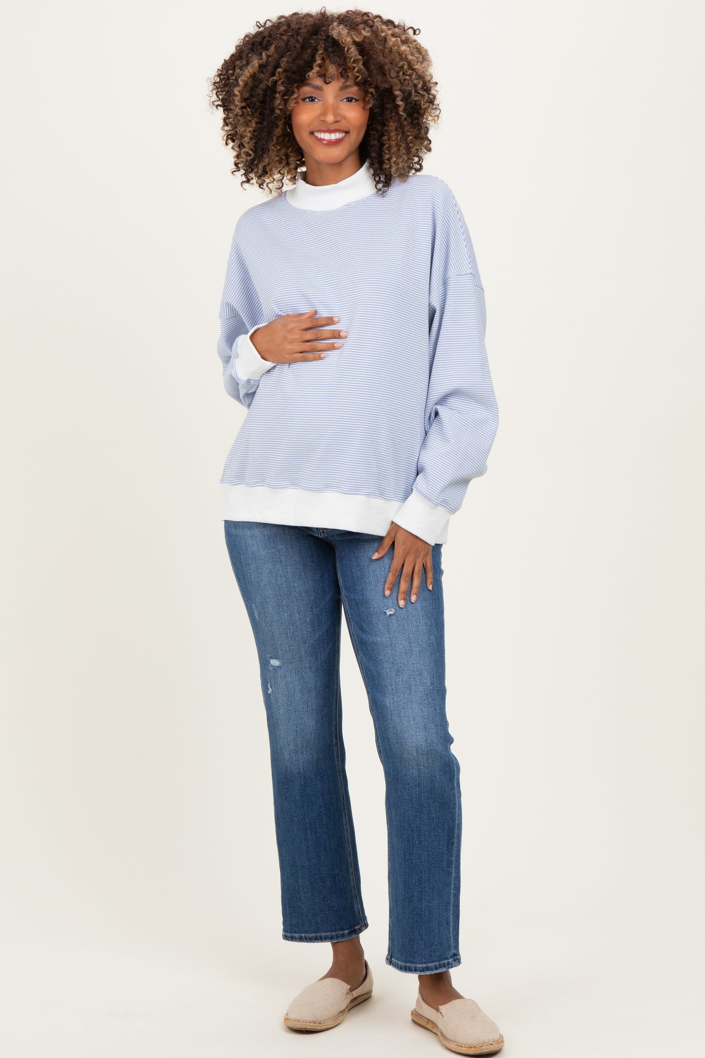woman wearing a light blue maternity long sleeve sweatshirt with a white mock neck and holding her baby bump, paired with jeans
