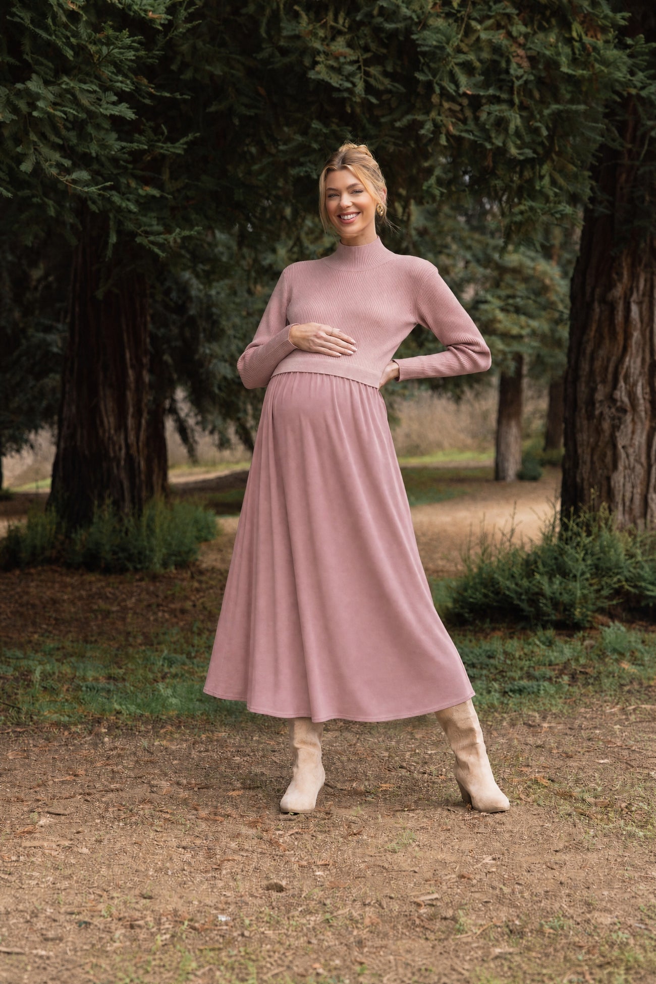 woman wearing a light pink maternity dress with a ribbed top and pleated skirt, holding her baby bump in an outdoor setting