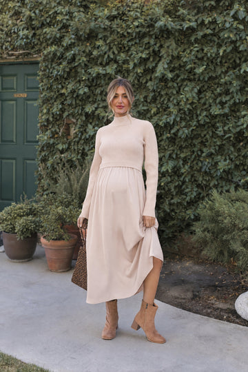 woman wearing a beige maternity sweater and a flowy beige maternity dress, holding a brown handbag and standing outdoors
