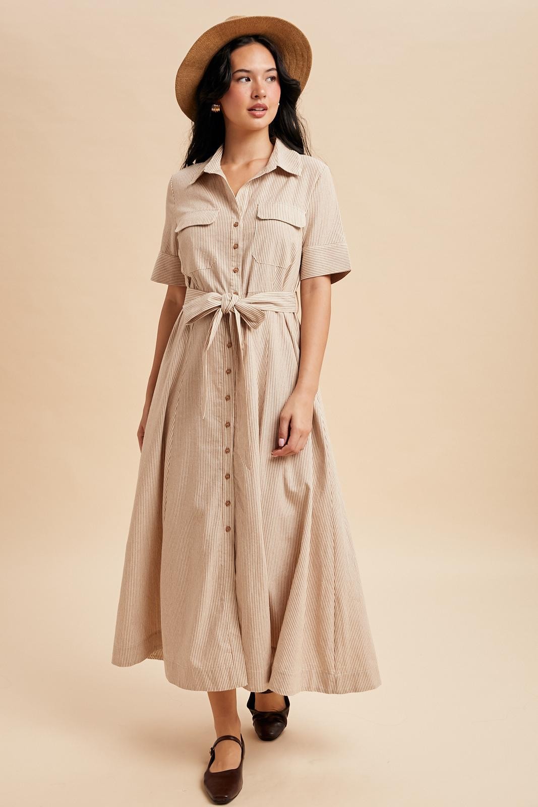 woman wearing a beige striped button-up maxi dress with a tie waist and short sleeves, paired with a hat and flats