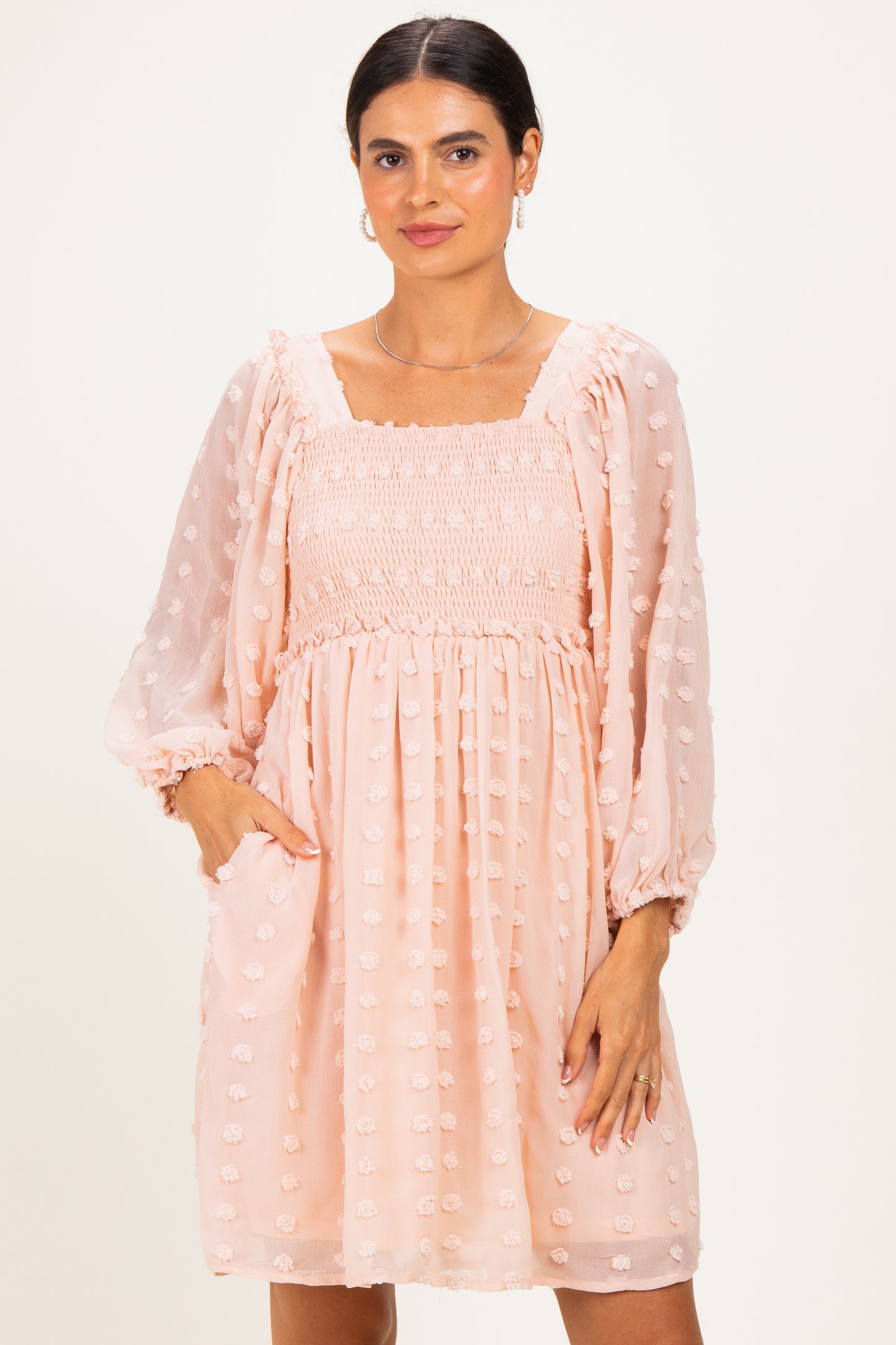 woman wearing a peach smocked dress with long sleeves and textured dots, standing with hands in pockets