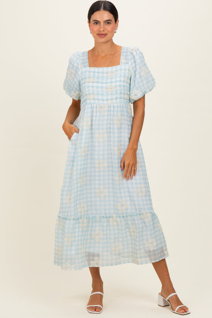 woman wearing a light blue gingham midi dress with puff sleeves and floral details, standing with hands in pockets