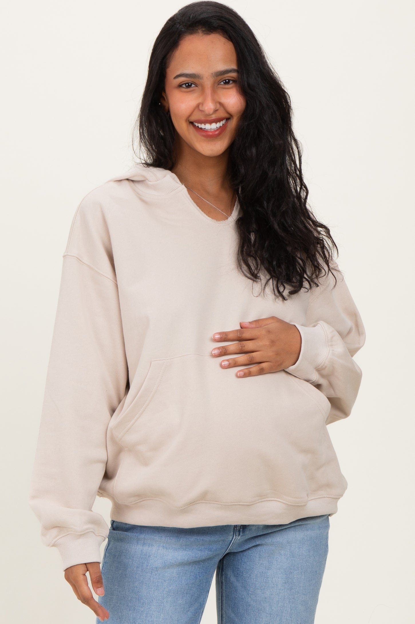 woman wearing a beige maternity hoodie and holding her baby bump while smiling