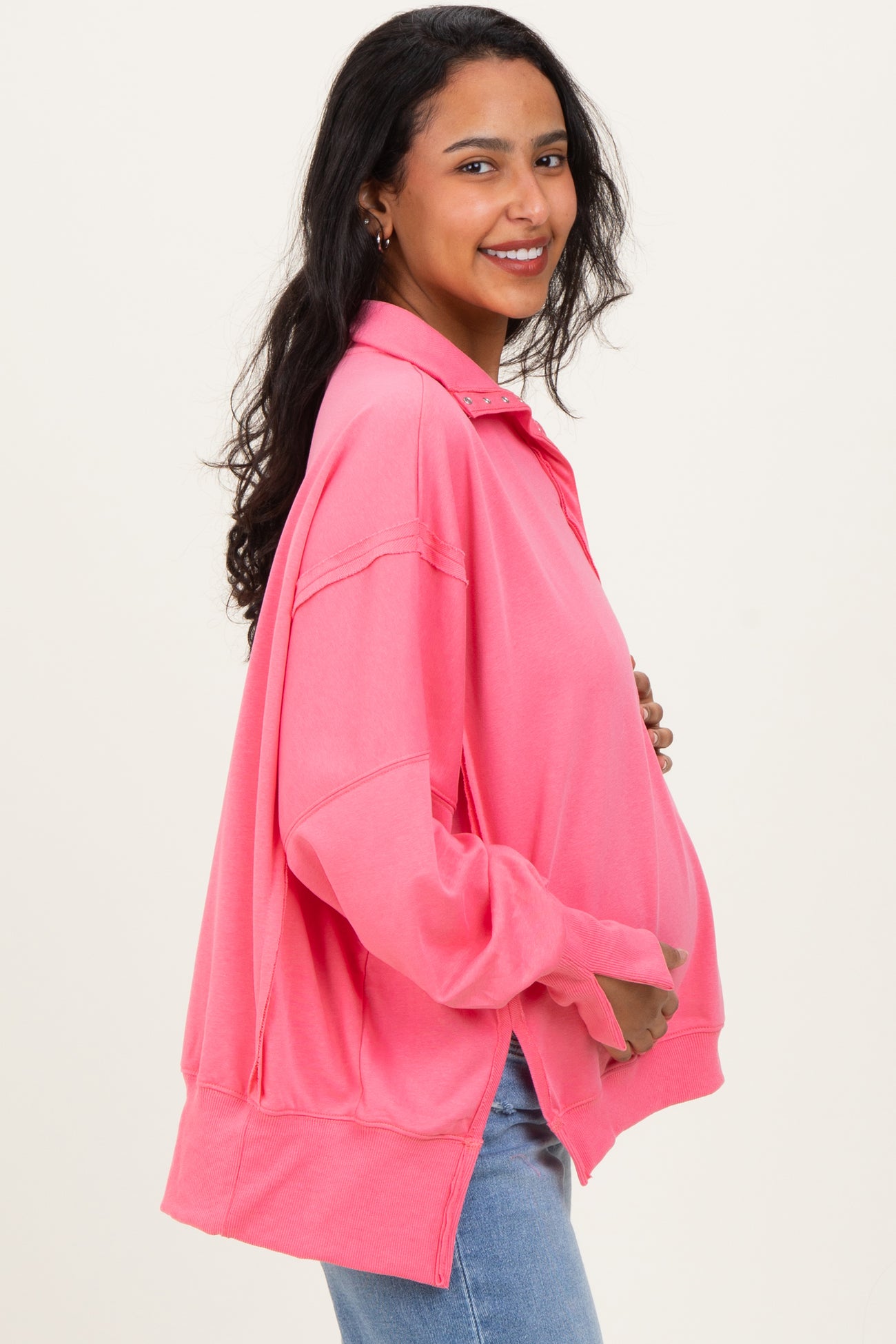 woman wearing a bright pink oversized maternity pullover, holding her baby bump, styled with jeans
