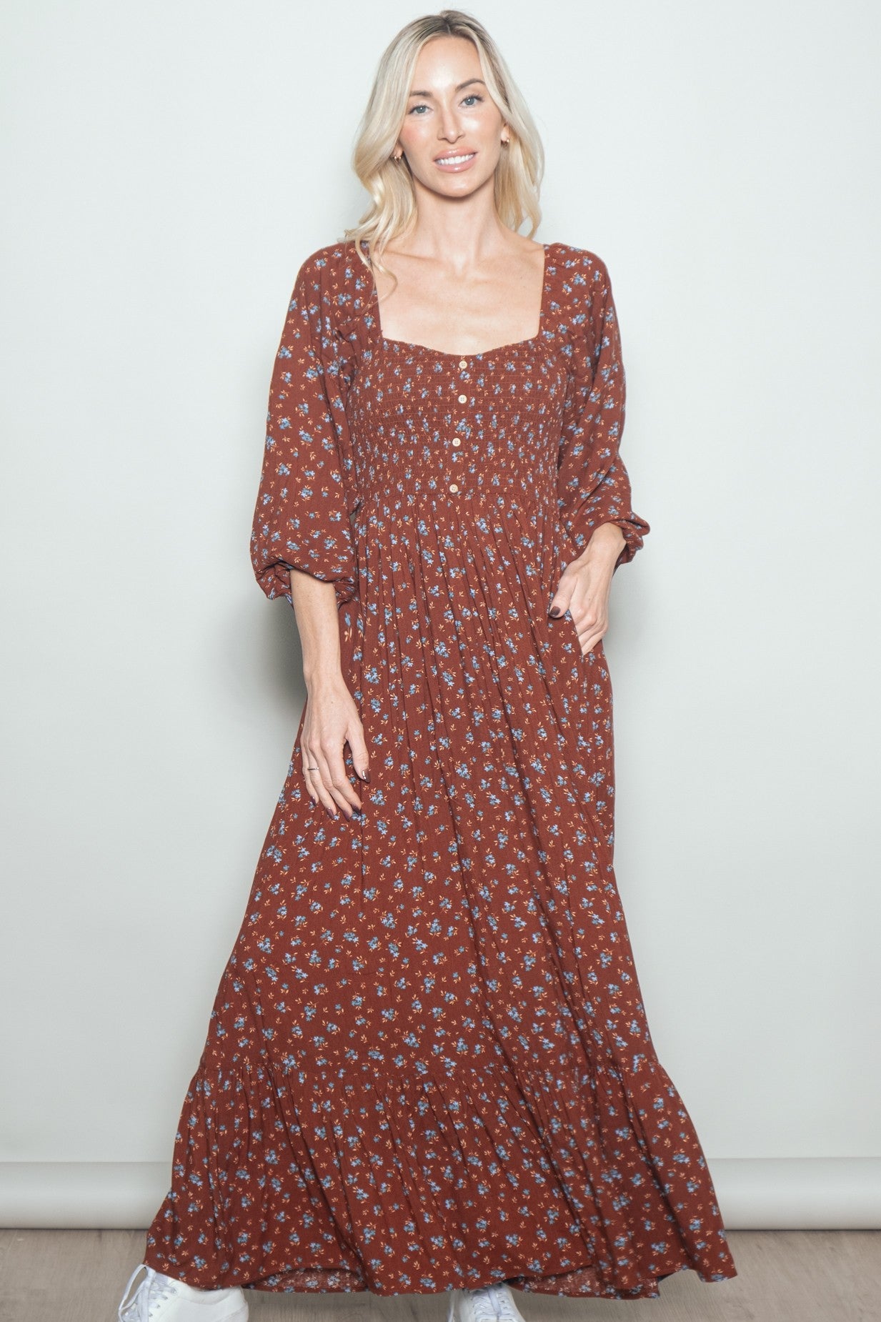 woman wearing a rust floral maxi dress with a square neckline and puff sleeves, standing with one hand in the pocket