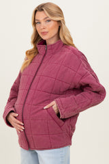 woman wearing a burgundy quilted maternity jacket with a standing collar and side pockets, gently cradling her baby bump
