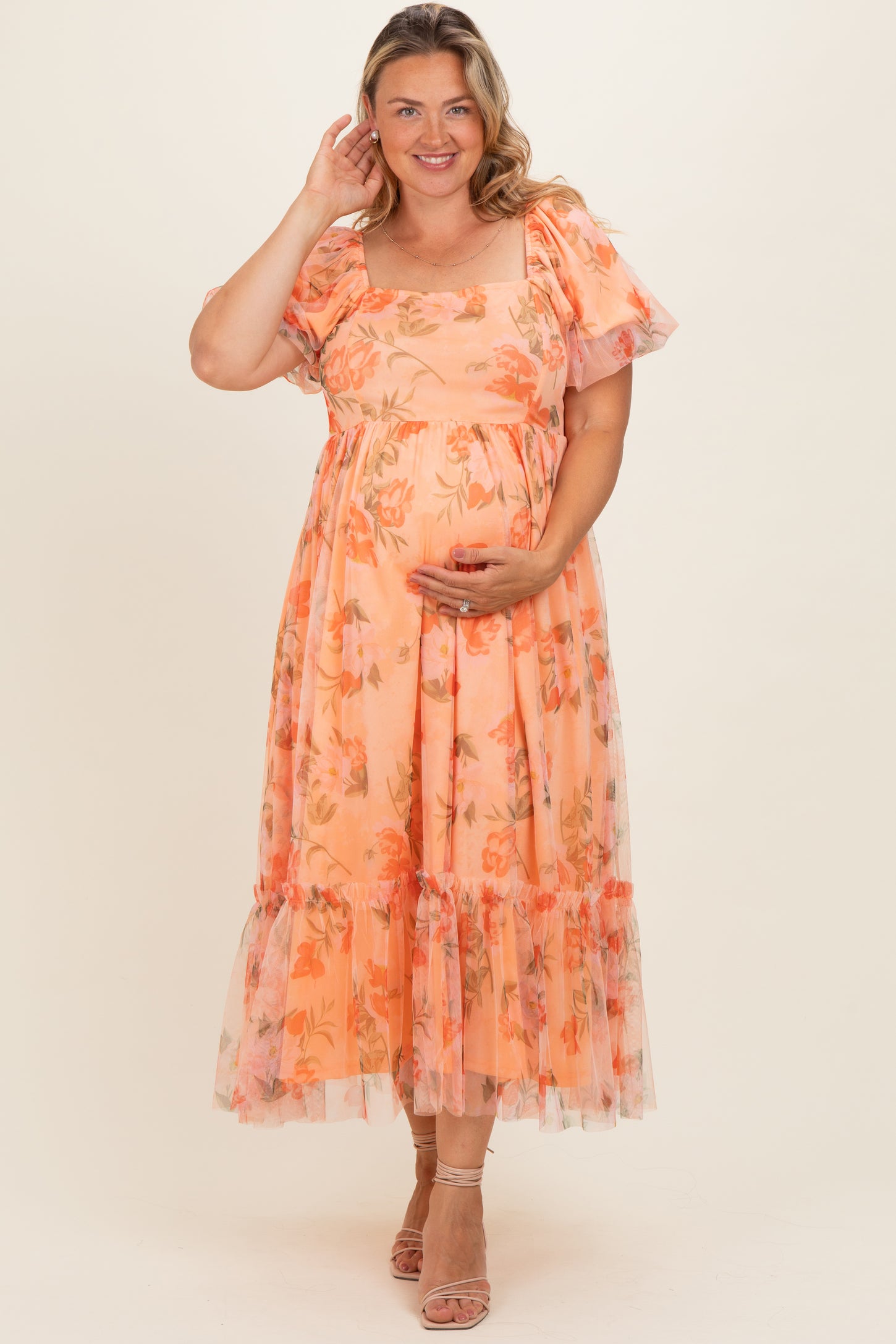 woman in a flowy peach floral maternity dress with puff sleeves and ruffled hem, gently holding her baby bump