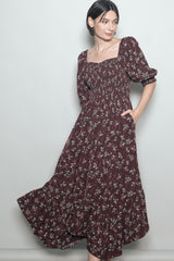 woman wearing a burgundy floral maxi dress with puff sleeves and a ruffled hem, posing with one hand in pocket