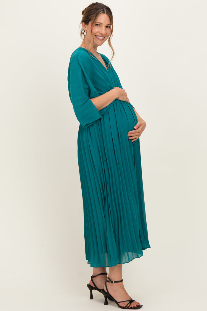 woman wearing a teal pleated maternity dress, holding her baby bump, standing in profile with a smile