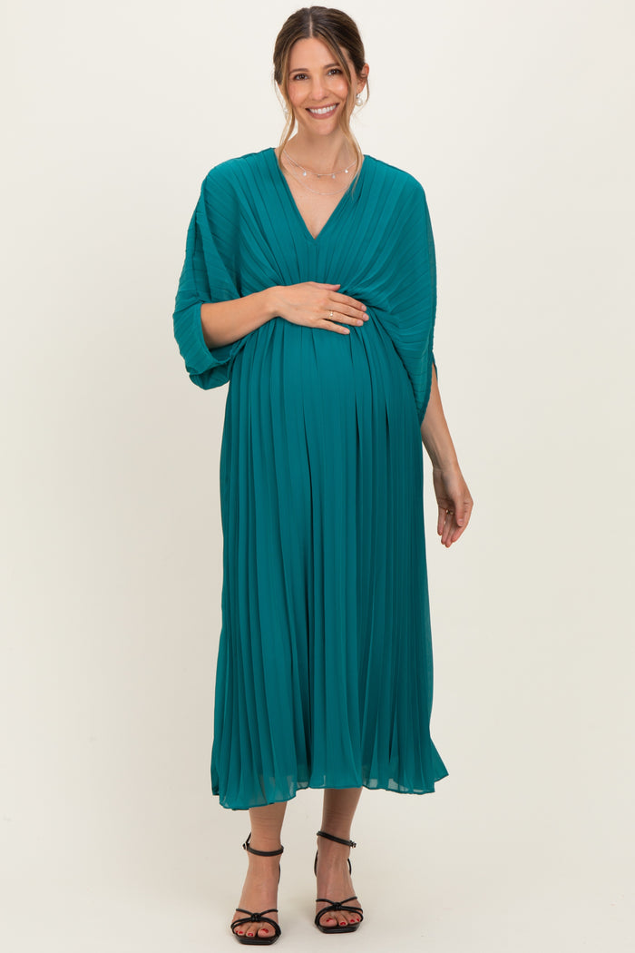 woman wearing a teal pleated maternity dress, gently cradling her baby bump