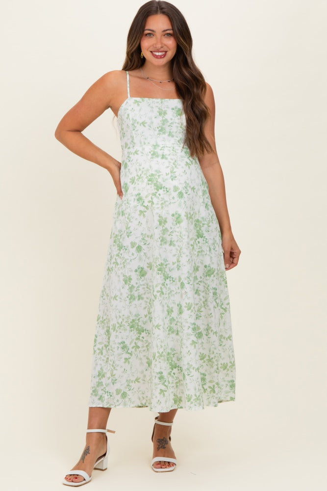 woman wearing a white floral maternity maxi dress with green details and spaghetti straps, posing with a smile