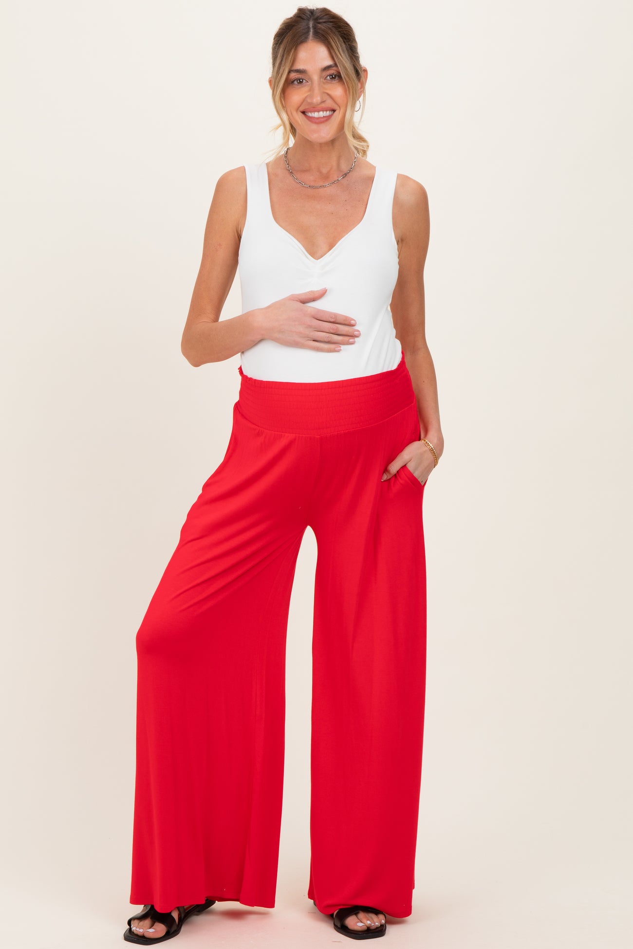 woman wearing a white sleeveless maternity top and red wide-leg maternity pants, holding her baby bump