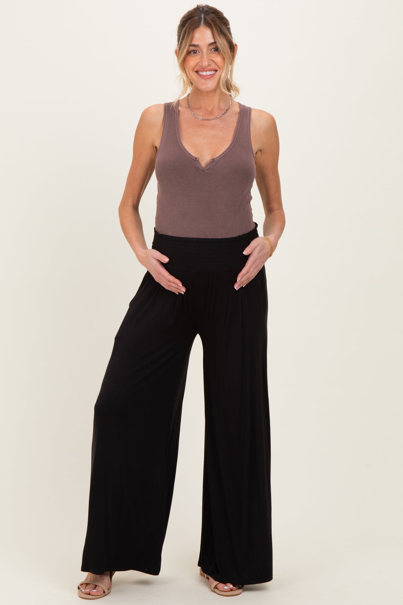 woman wearing a brown ribbed maternity tank top and black wide-leg maternity pants, holding her baby bump