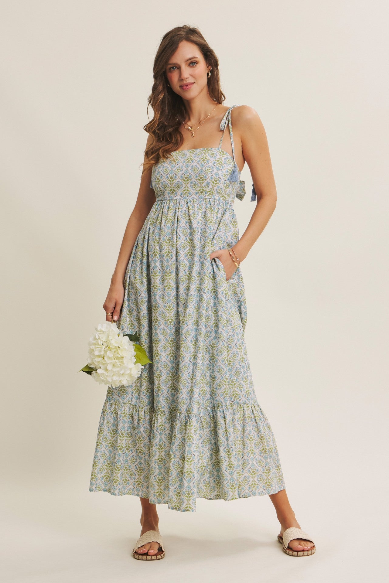 woman wearing a light blue floral maxi dress with tie straps and pockets, holding a bouquet of flowers